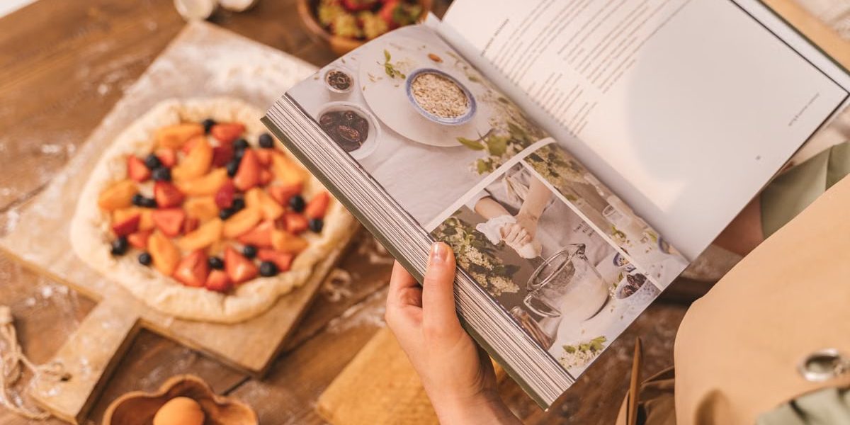 A person reads a cookbook while preparing a fruit tart on a wooden table.
