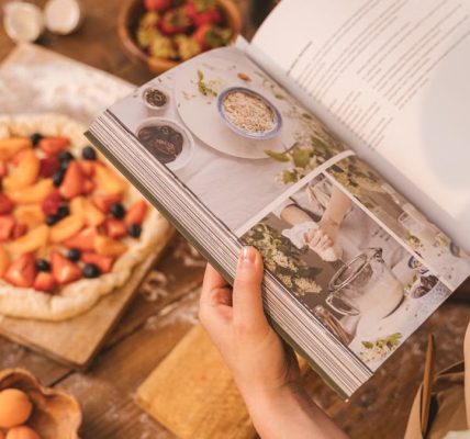 A person reads a cookbook while preparing a fruit tart on a wooden table.